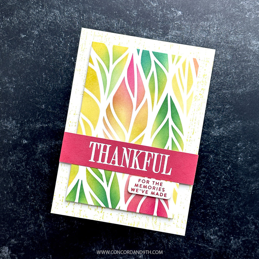A handmade greeting card with a colorful watercolor leaf pattern in yellow, green, and pink, overlaid with a red banner that reads 'THANKFUL'. The card is photographed flat on a dark, textured background.