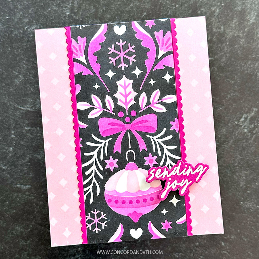 A handmade greeting card featuring a winter holiday design with pink and black patterns, a pink ornament, and a die-cut sticker that reads 'sending joy' on a dark textured background.