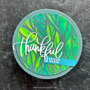A round handmade greeting card or coaster featuring a green and yellow leaf pattern, a gray circular border, and the word 'thankful' in white script. A small blue banner reads 'BEYOND WORDS'.