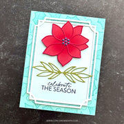 A handmade greeting card featuring a red poinsettia flower with green leaves and the text 'celebrate THE SEASON' on a light blue card, photographed on a dark textured surface.