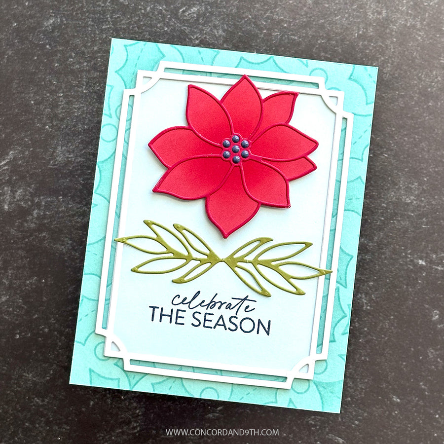 A handmade greeting card featuring a red poinsettia flower with green leaves and the text 'celebrate THE SEASON' on a light blue card, photographed on a dark textured surface.