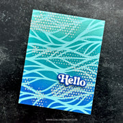 A handmade greeting card with a blue and teal wavy pattern, decorated with small silver glitter dots and a raised blue 'Hello' badge, photographed on a dark textured background.