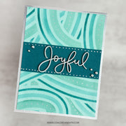 A handmade greeting card with a teal and mint green abstract wavy pattern, featuring a dark teal banner across the center with the word 'Joyful' in white script and small white dots. The card is photographed at an angle against a light gray marble background.