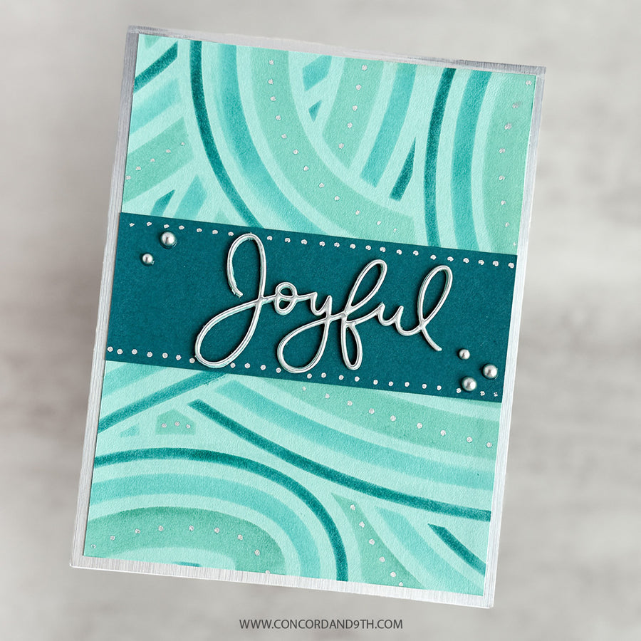 A handmade greeting card with a teal and mint green abstract wavy pattern, featuring a dark teal banner across the center with the word 'Joyful' in white script and small white dots. The card is photographed at an angle against a light gray marble background.