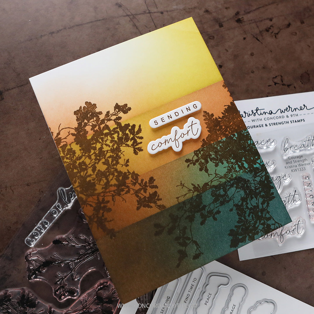 A handmade greeting card with a yellow-to-teal gradient background, dark tree silhouette overlays, and the words 'SENDING comfort' in layered die-cut stickers, displayed on a dark wood surface with stamp and embosser tools nearby.
