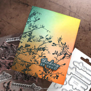 A handmade greeting card with a yellow-to-orange gradient background and black branch silhouette stamping, displayed with clear and metal embosser dies on a brown textured surface.