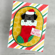 A handmade Christmas greeting card featuring a cartoon black puppy wearing a Santa hat peeking out of a red stocking with green trim. The card has a yellow oval center with gold polka dots, a red and green striped border, and a small white tag that reads 'HAPPY PAWIDAYS'.