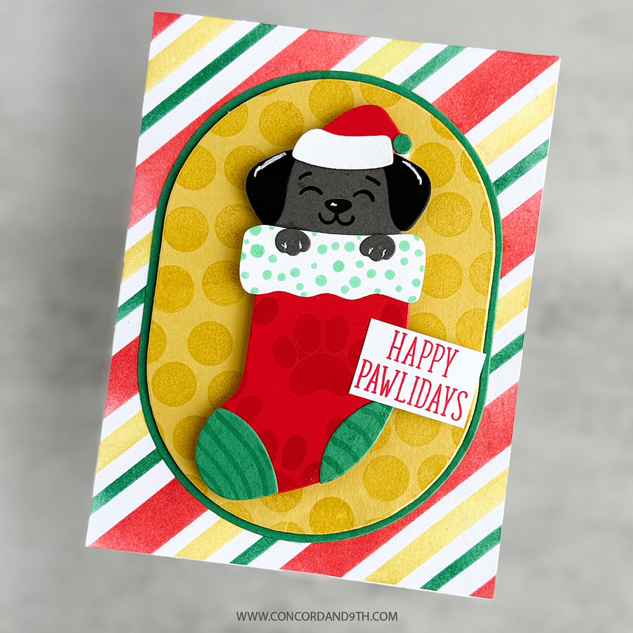 A handmade Christmas greeting card featuring a cartoon black puppy wearing a Santa hat peeking out of a red stocking with green trim. The card has a yellow oval center with gold polka dots, a red and green striped border, and a small white tag that reads 'HAPPY PAWIDAYS'.