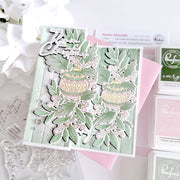 A handmade layered card featuring a Christmas tree design in sage green, cream, and pink, styled with clear stamps and pink ink pads on a white background.