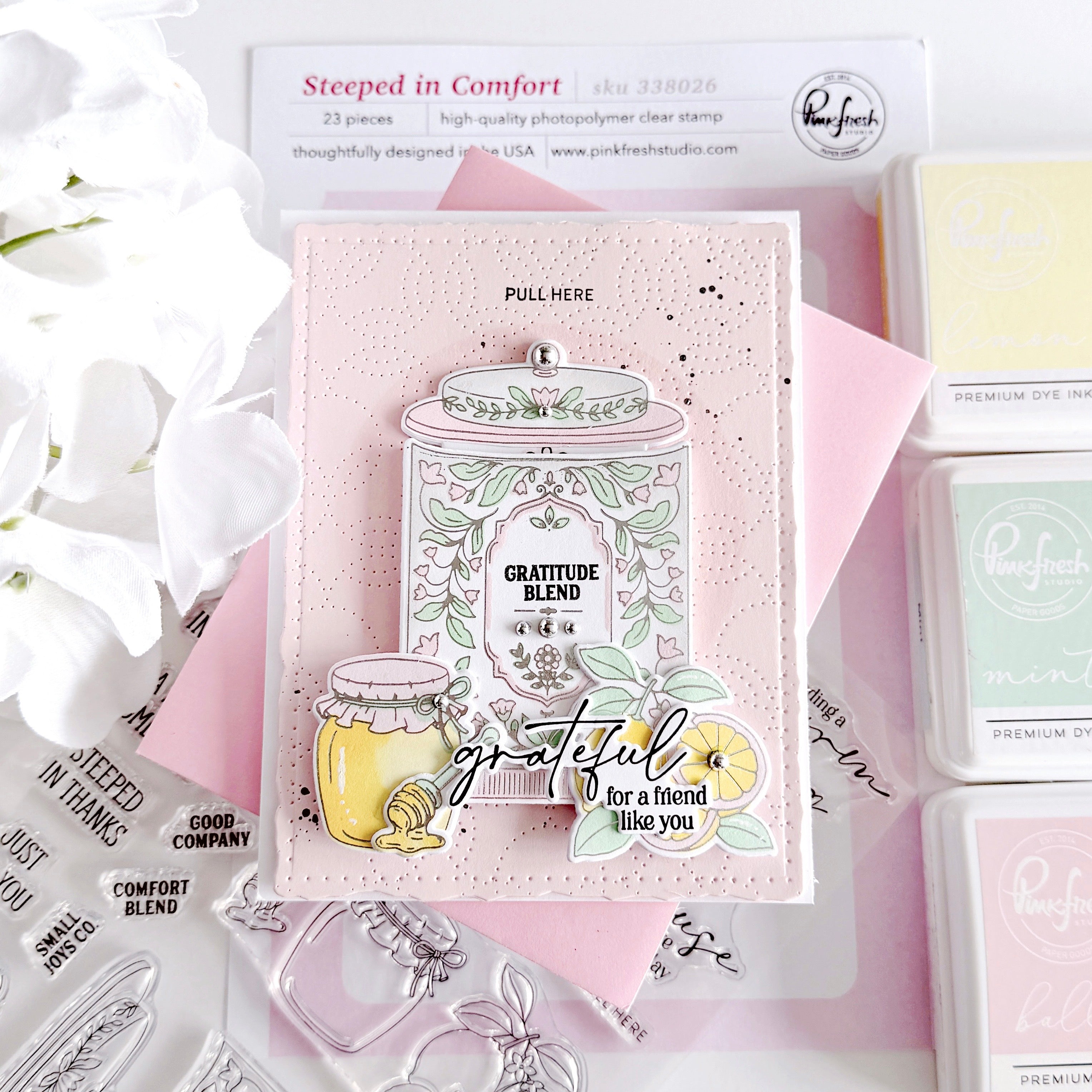 A handmade layered paper craft stamp card featuring a jar of honey with the words 'Gratitude Blend' and 'grateful for a friend like you', styled on a pink background with other crafting supplies.