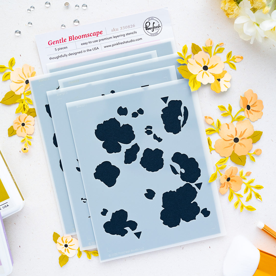 A flat lay of layered stencils, surrounded by yellow and peach paper flowers on a white surface. A card at the top rwith brand Pinkfresh Studio reads 'Gentle Bloomscape' with product details, and small craft tools are partially visible around the edges.