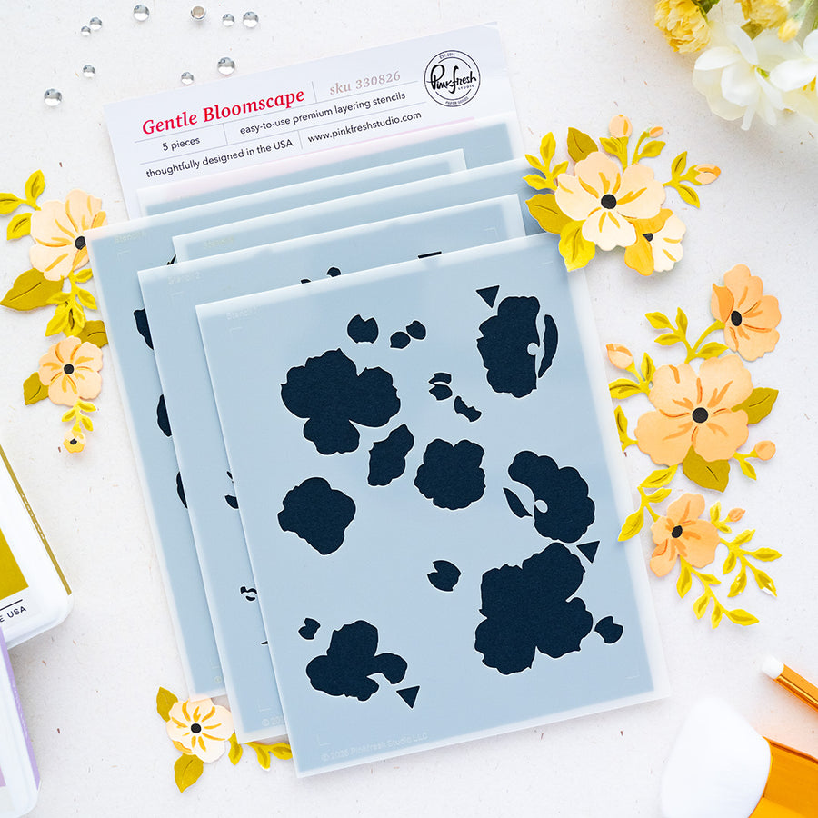 A flat lay of layered stencils, surrounded by yellow and peach paper flowers on a white surface. A card at the top rwith brand Pinkfresh Studio reads 'Gentle Bloomscape' with product details, and small craft tools are partially visible around the edges.