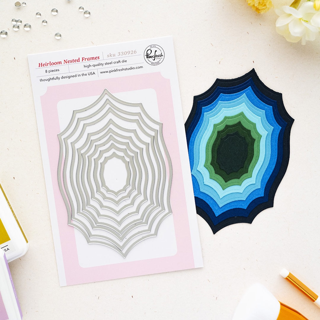 A flat lay of a steel card die set on a light cream surface. On the left is the printed package card for Pinkfresh Studio 'Heirloom Nested Frames' showing a silver nested spiderweb-style die, and on the right is the corresponding layered steel cutout in shades of blue, green, and black.