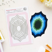 A flat lay of a steel card die set on a light cream surface. On the left is the printed package card for Pinkfresh Studio 'Heirloom Nested Frames' showing a silver nested spiderweb-style die, and on the right is the corresponding layered steel cutout in shades of blue, green, and black.