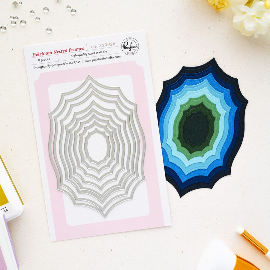 A flat lay of a steel card die set on a light cream surface. On the left is the printed package card for Pinkfresh Studio 'Heirloom Nested Frames' showing a silver nested spiderweb-style die, and on the right is the corresponding layered steel cutout in shades of blue, green, and black.