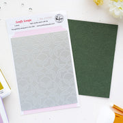 A steel craft die set by Pinkfresh Studio called 'Leafy Loops' shown with its packaging card and a dark green textured sheet of paper on a white flat-lay surface.