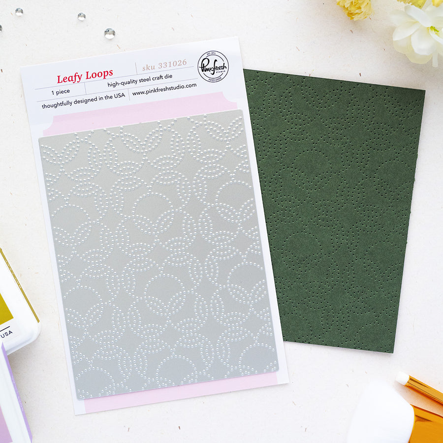 A steel craft die set by Pinkfresh Studio called 'Leafy Loops' shown with its packaging card and a dark green textured sheet of paper on a white flat-lay surface.