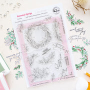 A sheet of clear photopolymer stamps titled 'Seasonal Sprigs' by Pinkfresh Studio, displayed on a light beige surface with decorative floral and script elements. The sheet contains nine clear stamps with winter wreath and holiday message designs over a pale pink backing card.