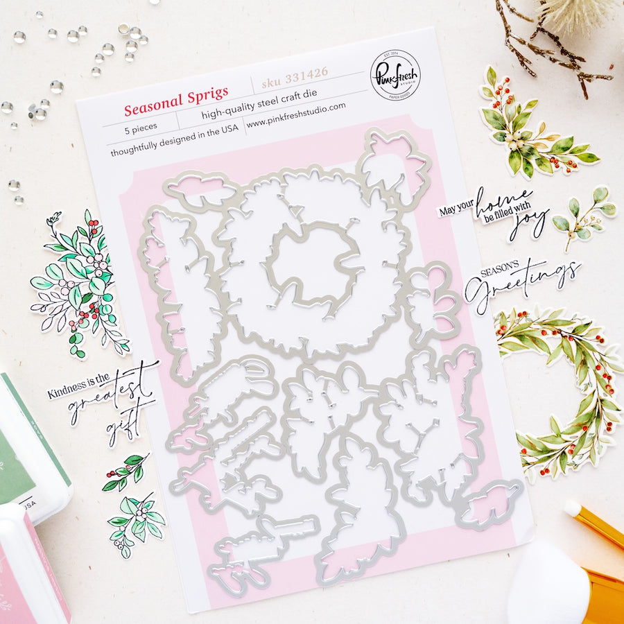 A flat-lay product photo of a Steel Cut die set by Pinkfresh Studio, titled 'Seasonal Sprigs'. The set includes 5 steel dies on a white backing card with pink borders, surrounded by styled craft props including stamped botanicals, a pen, and a paintbrush on a white background.