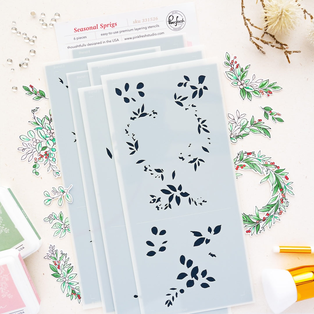 A set of layered stencils from the brand Pinkfresh Studio, shown in a styled flat-lay with coordinating floral stamps, ink pads, and craft tools on a white background.