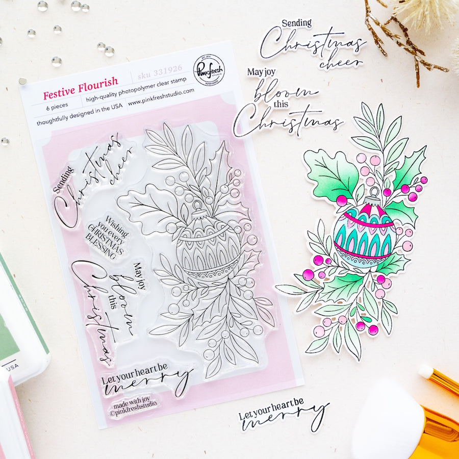 A flat-lay product photo of a clear stamp set called 'Festive Flourish' by Pinkfresh Studio, displayed on a white background with a pink backing card. The stamp design features a hanging basket with green leaves, pink berries, and black ink line art.