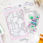 Festive Flourish steel craft die set by Pinkfresh Studio, shown with its packaging and a color sample illustration of a Christmas ornament and holly leaves on a light beige background.