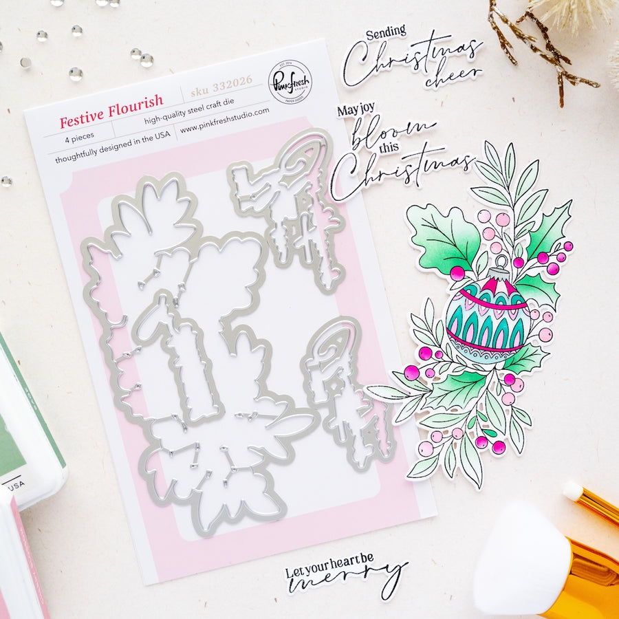 Festive Flourish steel craft die set by Pinkfresh Studio, shown with its packaging and a color sample illustration of a Christmas ornament and holly leaves on a light beige background.