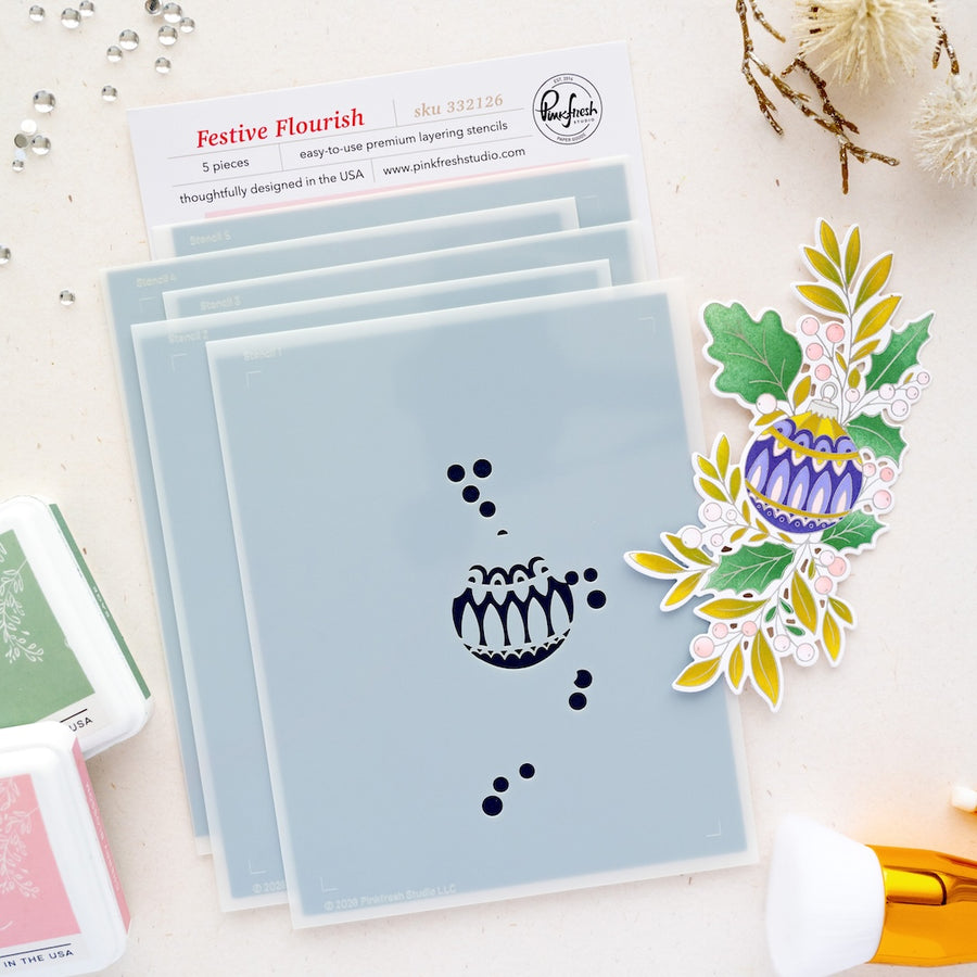 A stack of Festive Flourish premium layered stencils by Pinkfresh Studio is displayed on a light beige surface with craft supplies. The stencils feature a winter berry design, and a colorful stamped floral berry cutout is shown to the right.