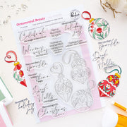 A clear stamp set and matching stamped holiday ornament designs displayed on a white flat-lay background with decorative Christmas-themed props. The sheet is labeled 'Ornamental Beauty' and shows 11 clear stamps for crafting holiday phrases and ornament shapes by Pinkfresh Studio