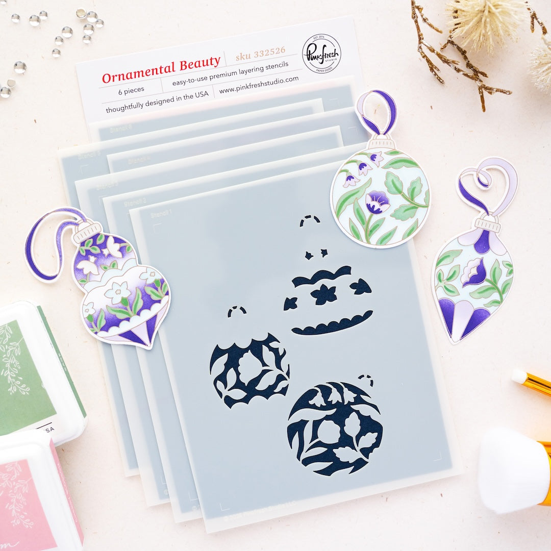 A flat-lay of a craft stencils labeled 'Ornamental Beauty' by Pinkfresh Studio with six layered stencil sheets and three matching die-cut ornament stencils featuring purple, green, and white floral patterns on a light pink background.