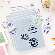 A flat-lay of a craft stencils labeled 'Ornamental Beauty' by Pinkfresh Studio with six layered stencil sheets and three matching die-cut ornament stencils featuring purple, green, and white floral patterns on a light pink background.