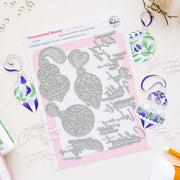 A flat-lay of a metal press plate set by Ornamental Beauty by Pinkfresh Studio, featuring Christmas ornament and script designs, surrounded by colorful stamped and painted ornament samples on a light beige surface.