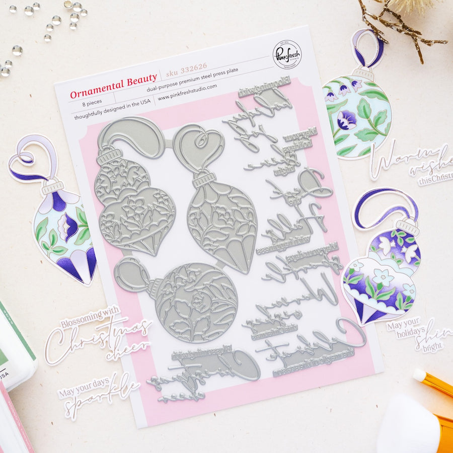 A flat-lay of a metal press plate set by Ornamental Beauty by Pinkfresh Studio, featuring Christmas ornament and script designs, surrounded by colorful stamped and painted ornament samples on a light beige surface.