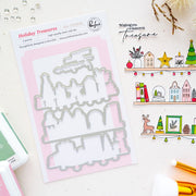 A flat-lay product photo of a 'Holiday Treasures' 2-piece metal die set by Pinkfresh Studio. The die set is packaged in a white card with a pink background, displayed next to coordinating stamped and stamped-out Christmas illustrations on a light cream surface.
