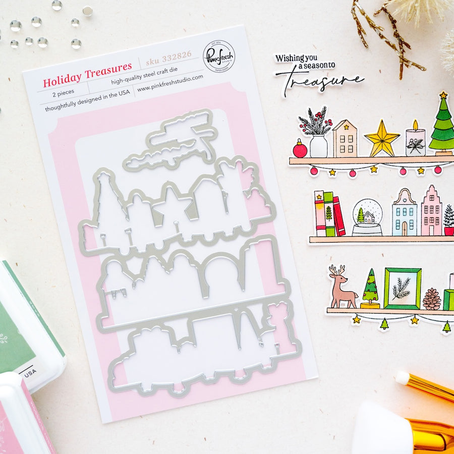 A flat-lay product photo of a 'Holiday Treasures' 2-piece metal die set by Pinkfresh Studio. The die set is packaged in a white card with a pink background, displayed next to coordinating stamped and stamped-out Christmas illustrations on a light cream surface.