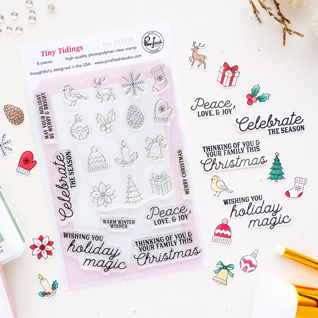 A sheet of clear stamp designs called Tiny Tidings by Pinkfresh Studio featuring Christmas-themed illustrations and phrases like 'Peace, Love, & Joy' and 'Celebrate the Season,' shown on a light background with small decorative props.
