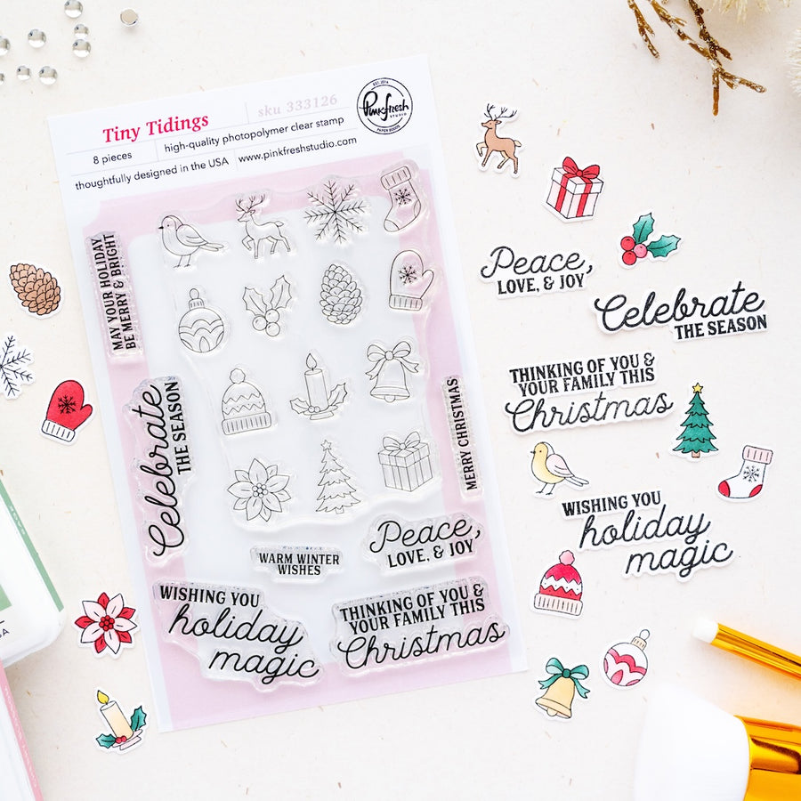 A sheet of clear stamp designs called Tiny Tidings by Pinkfresh Studio featuring Christmas-themed illustrations and phrases like 'Peace, Love, & Joy' and 'Celebrate the Season,' shown on a light background with small decorative props.