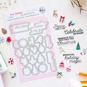 A flat-lay of a Christmas-themed die set called Tiny Tidings by Pinkfresh Studio, featuring a sheet of 5-piece steel dies on a pink backing card, surrounded by coordinating stamped and cut holiday designs on a white background.