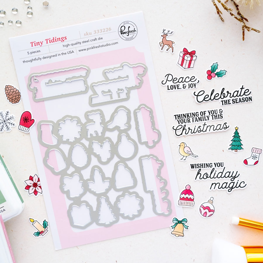 A flat-lay of a Christmas-themed die set called Tiny Tidings by Pinkfresh Studio, featuring a sheet of 5-piece steel dies on a pink backing card, surrounded by coordinating stamped and cut holiday designs on a white background.