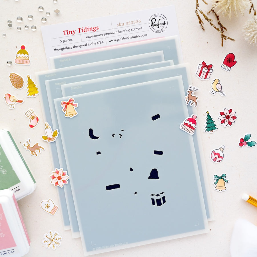 A flat-lay of stencils labeled 'Tiny Tidings' by Pinkfresh Studio small Christmas-themed die cuts and embellishments on a white surface.
