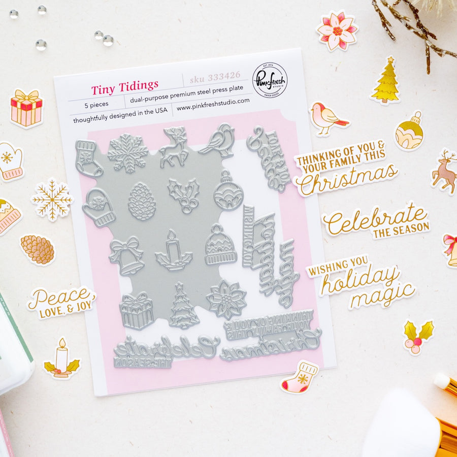 A flat-lay product photo of a 'Tiny Tidings' 5-piece double-purpose steel press plate on a white background decorated with holiday-themed stickers and ephemera. The product consists of a white backing card with a pink border and a silver-colored steel plate featuring Christmas motifs like snowflakes, ornaments, and a Christmas tree. Brand is Pinkfresh Studio.