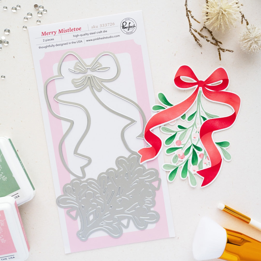 A holiday die set called Merry Mistletoe by brand Pinkfresh Studio on a pink backing card, styled on a white surface with decorative dried flowers, ink pads, and brushes. Pinkfresh Studio Merry Mistletoe Dies