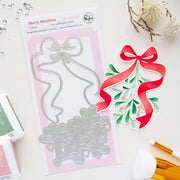 A holiday die set called Merry Mistletoe by brand Pinkfresh Studio on a pink backing card, styled on a white surface with decorative dried flowers, ink pads, and brushes. Pinkfresh Studio Merry Mistletoe Dies
