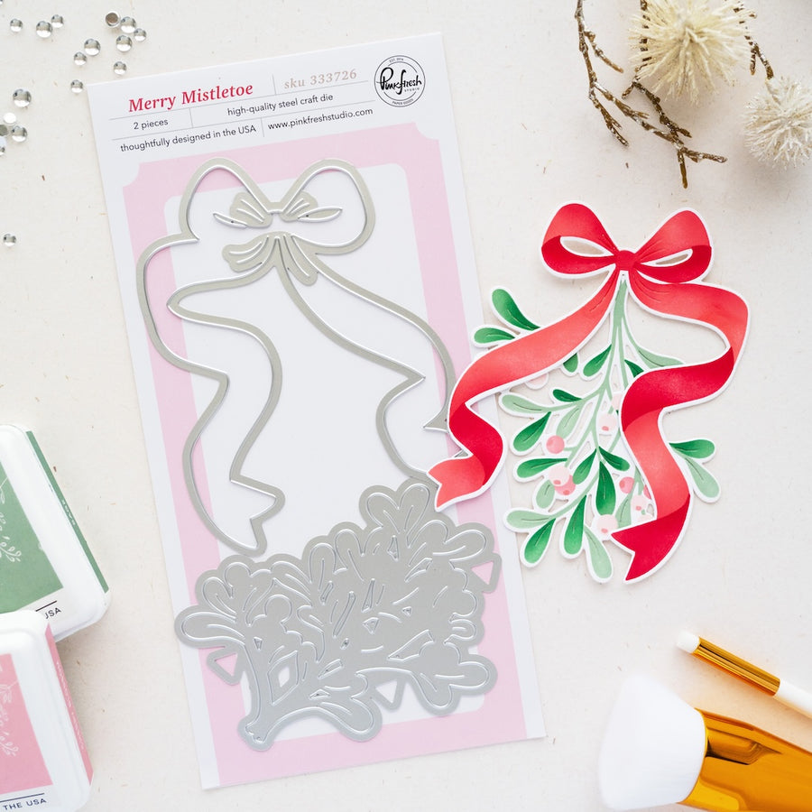 A holiday die set called Merry Mistletoe by brand Pinkfresh Studio on a pink backing card, styled on a white surface with decorative dried flowers, ink pads, and brushes. Pinkfresh Studio Merry Mistletoe Dies