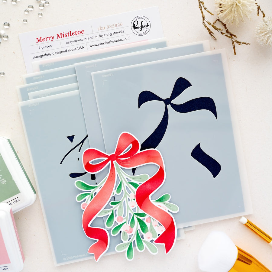 A flat-lay of layered stencils by Pinkfresh Studio. A white product card labeled 'Merry Mistletoe' are visible on a light beige background with small decorative props.