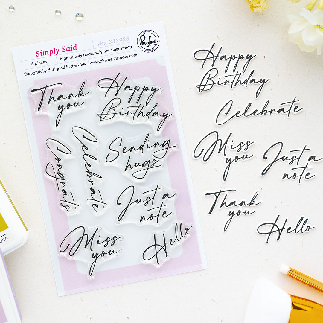 A flat-lay of Pinkfresh Studio's Simply Said 8-piece clear photopolymer stamp set on a white background, alongside matching black script letter stickers spelling birthday and greeting phrases.