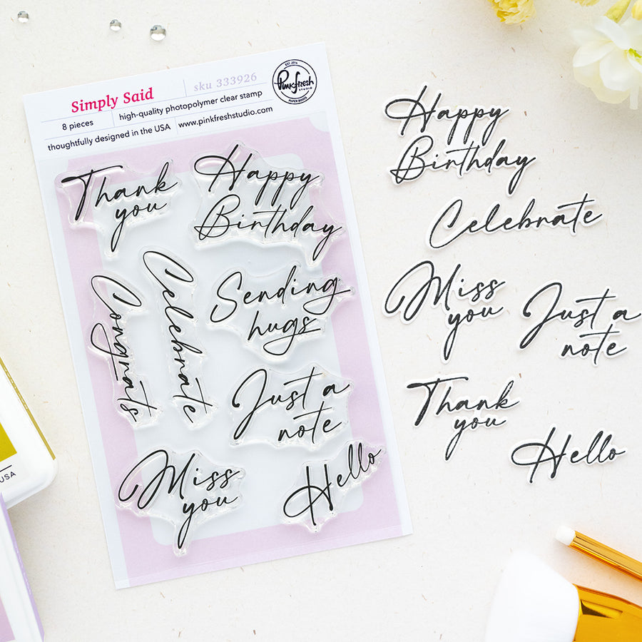 A flat-lay of Pinkfresh Studio's Simply Said 8-piece clear photopolymer stamp set on a white background, alongside matching black script letter stickers spelling birthday and greeting phrases.