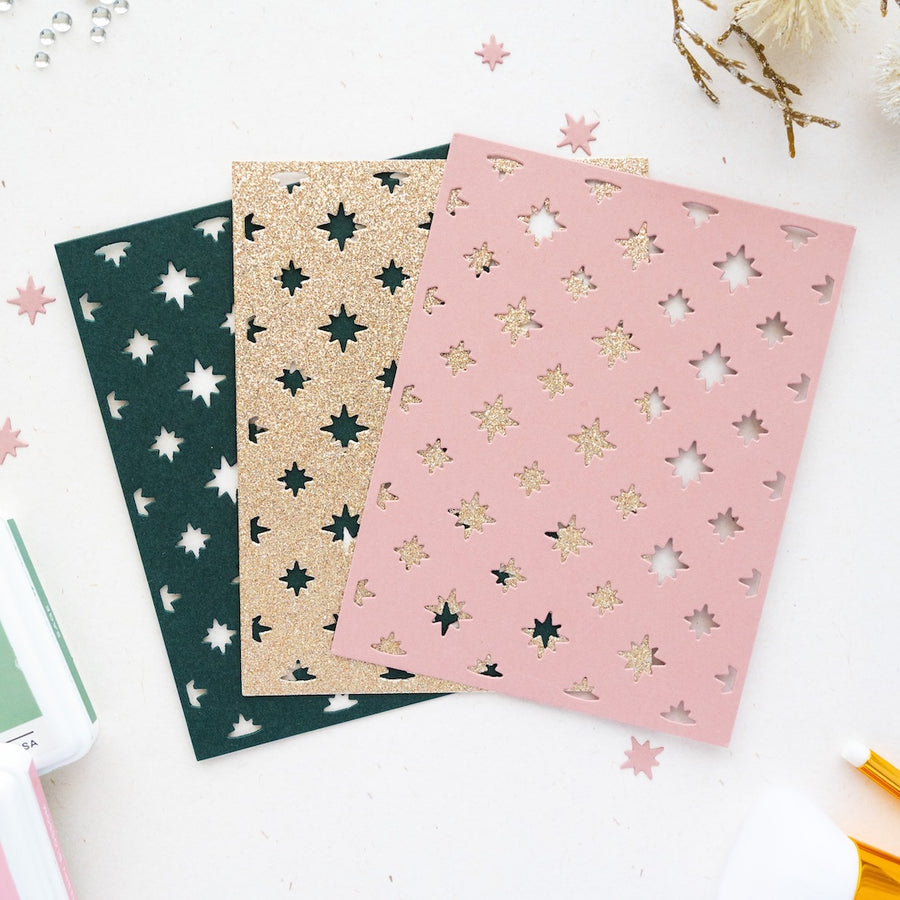 Three decorative die-cut cards or paper sheets with star and moon patterns, displayed in dark green, glittery gold, and dusty pink on a white background with scattered paper confetti.