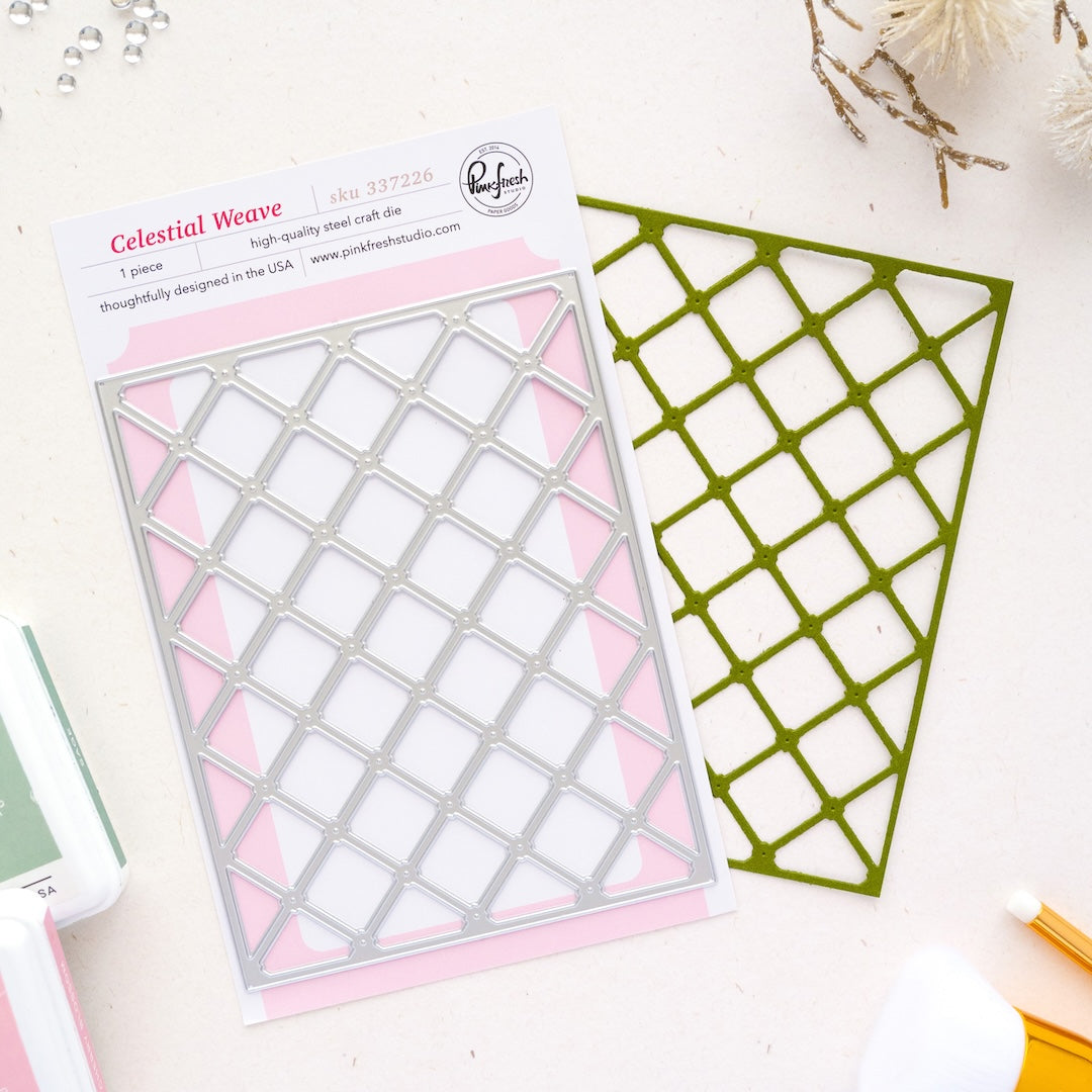A packaged metal die set labeled 'Celestial Weave' by Pinkfresh Studio, shown on a light surface with the die cutout and backing card visible. The product is photographed in a styled flat-lay with a few small craft props around it.