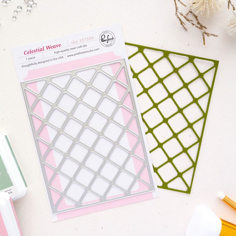 A packaged metal die set labeled 'Celestial Weave' by Pinkfresh Studio, shown on a light surface with the die cutout and backing card visible. The product is photographed in a styled flat-lay with a few small craft props around it.